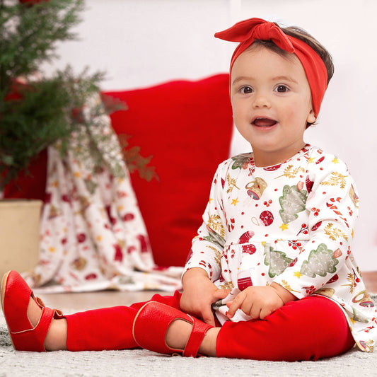 A baby girl  sitting in a room decorated for Christmas, wearing clothing that includes a Christmas-themed bamboo top and red leggings.