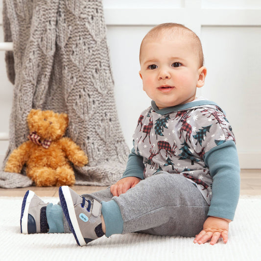 Baby sitting on a white surface with a teddy bear and knitted blanket in the background.