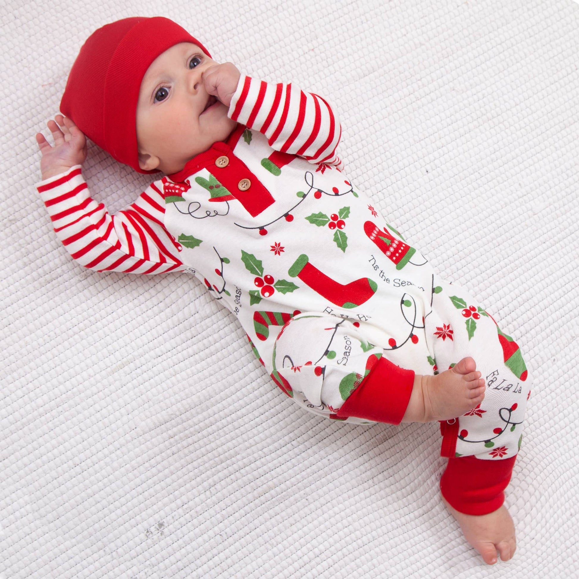 Baby wearing a red and white striped holiday-themed onesie and hat on a white textured surface.