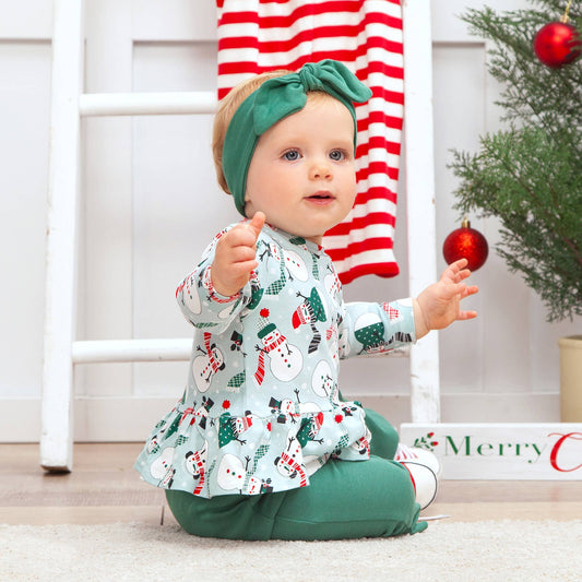 Baby in festive outfit with green headband and snowman patterned clothing.