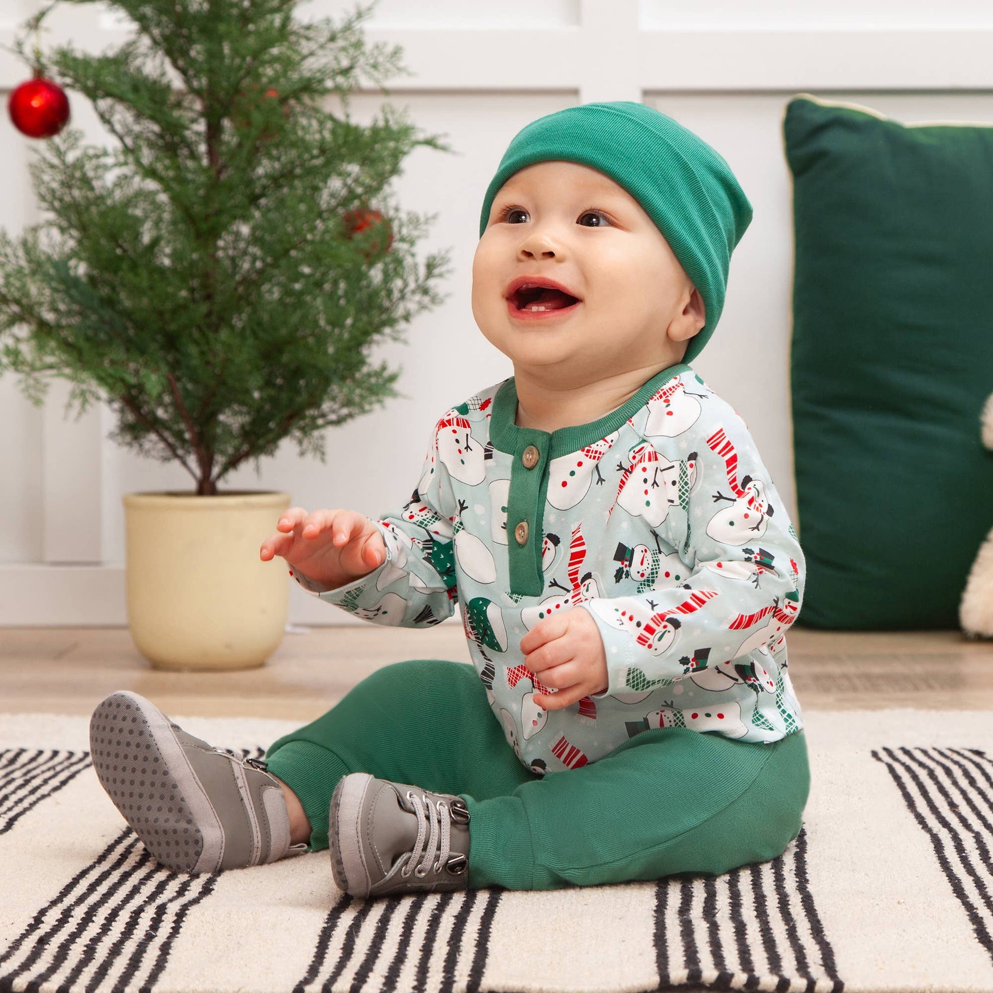 Baby in green hat and matching outfit sitting on a striped rug with a Christmas tree in the background.