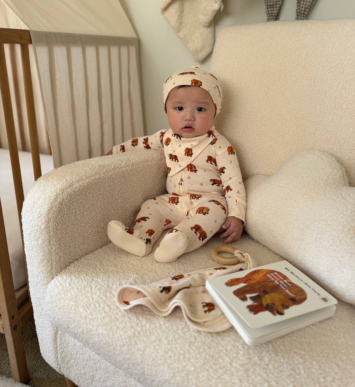 A baby wearing a cream-colored one-piece sleeper with brown bear prints and a matching beanie sits on a cream boucle armchair next to a wooden crib. A matching folded blanket and a baby book with a bear illustration are placed on the chair.