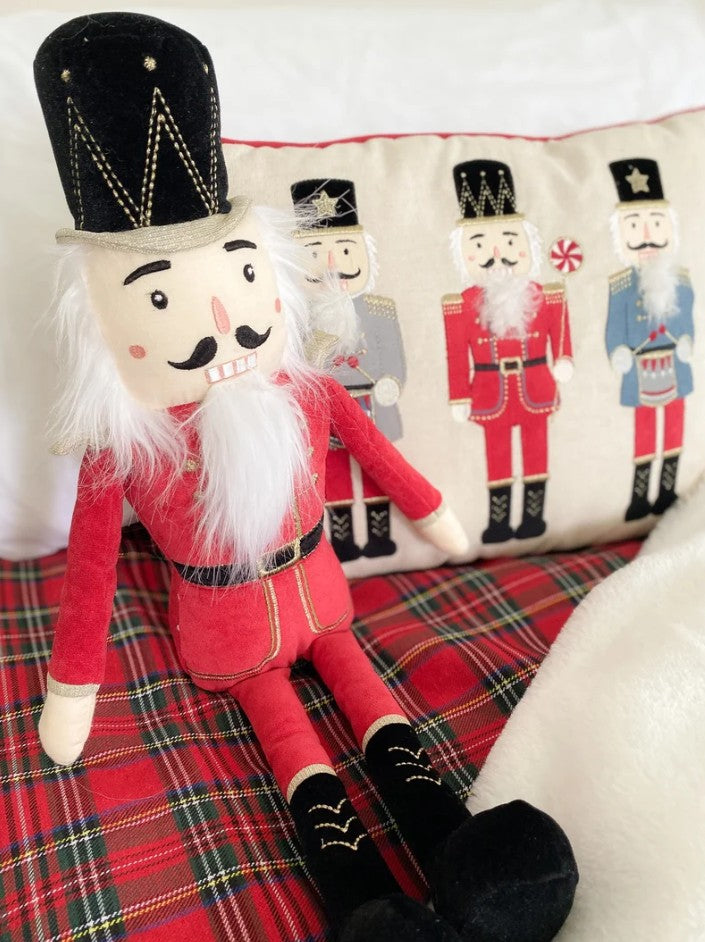 A plush nutcracker doll dressed in a red coat and black hat with white beard, posed on a red plaid blanket in front of a decorative pillow showing embroidered nutcracker figures.
