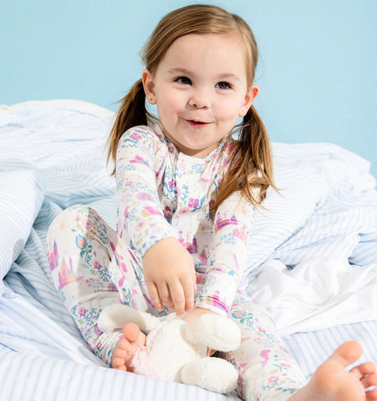 Child in castle and floral pajamas holding a teddy bear on a bed with a light blue background