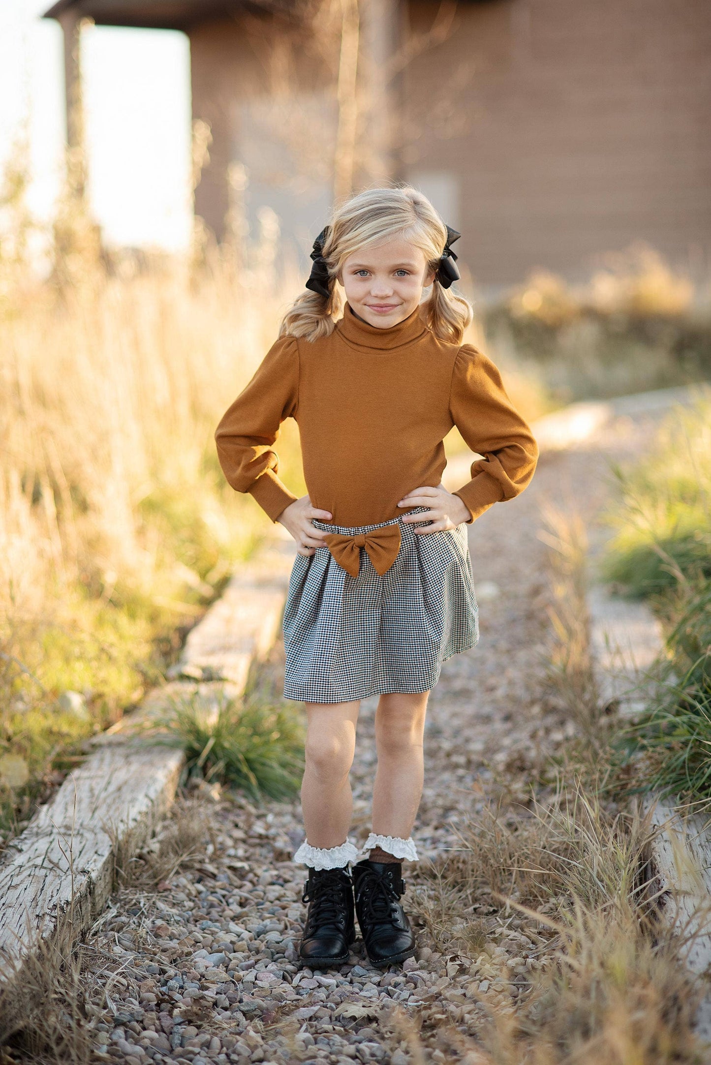 A girl wearing a caramel colored long puff sleeve top with a black and white houndstooth check pleated skirt with matching bow, standing on a gravel path.