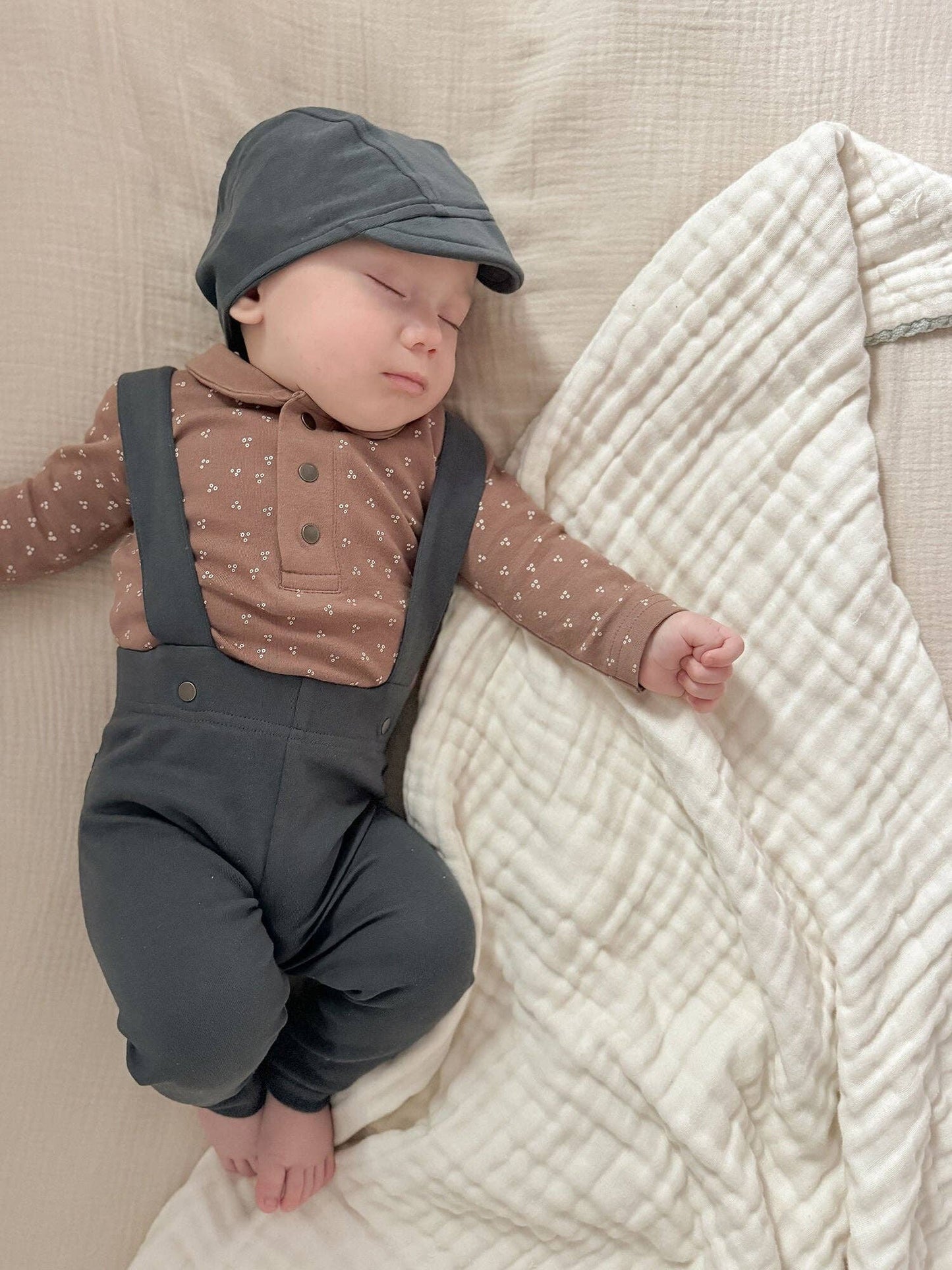 Baby wearing a brown shirt and charcoal pants and cap, sleeping on a beige blanket.