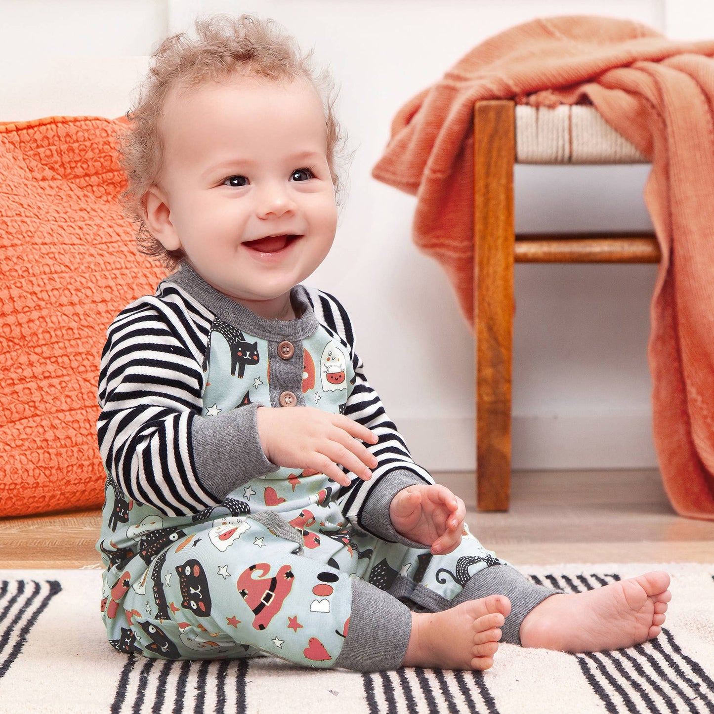 Baby sitting on a rug with a Halloween striped shirt and patterned pants in a home setting.