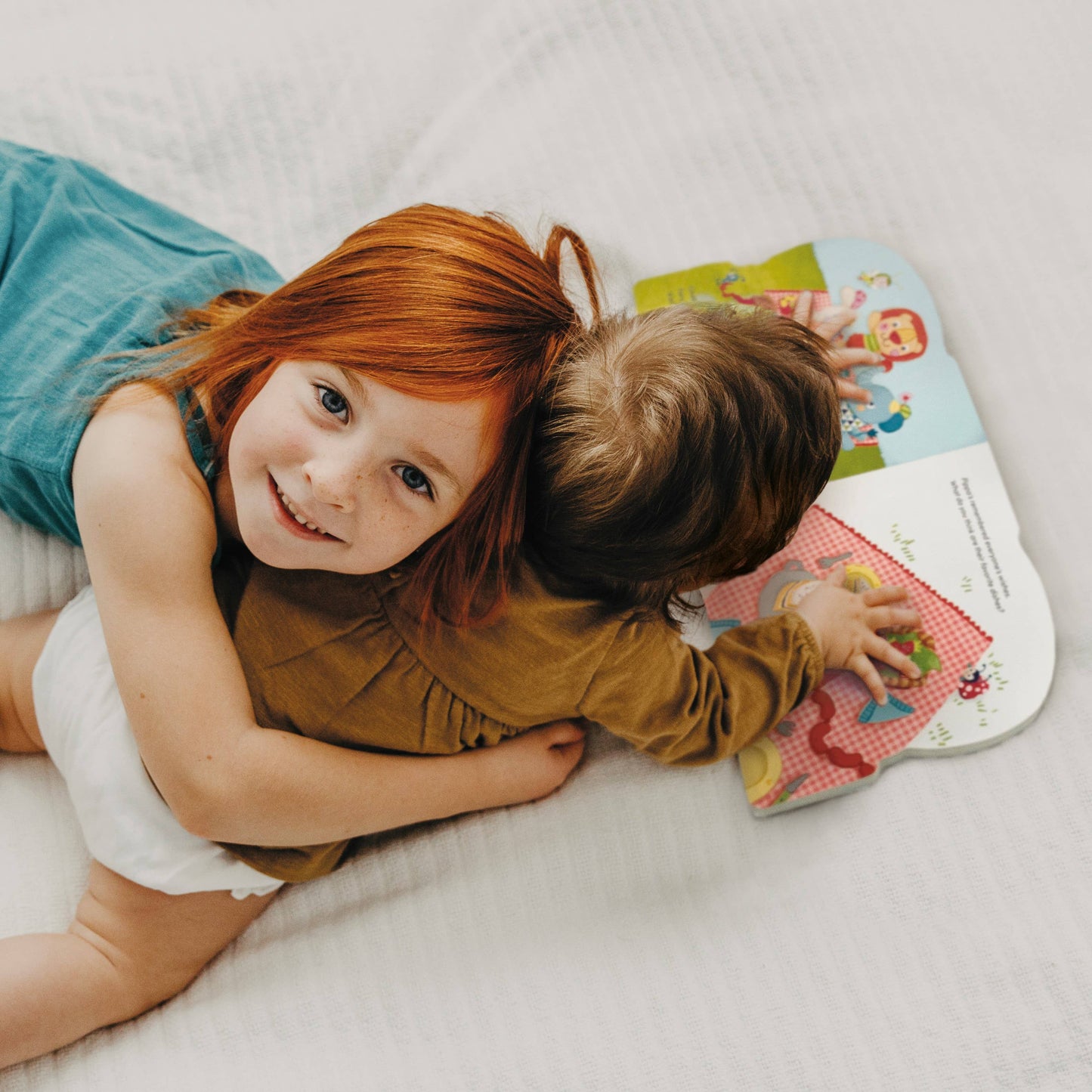 Two children hugging on a bed with a colorful book open between them.