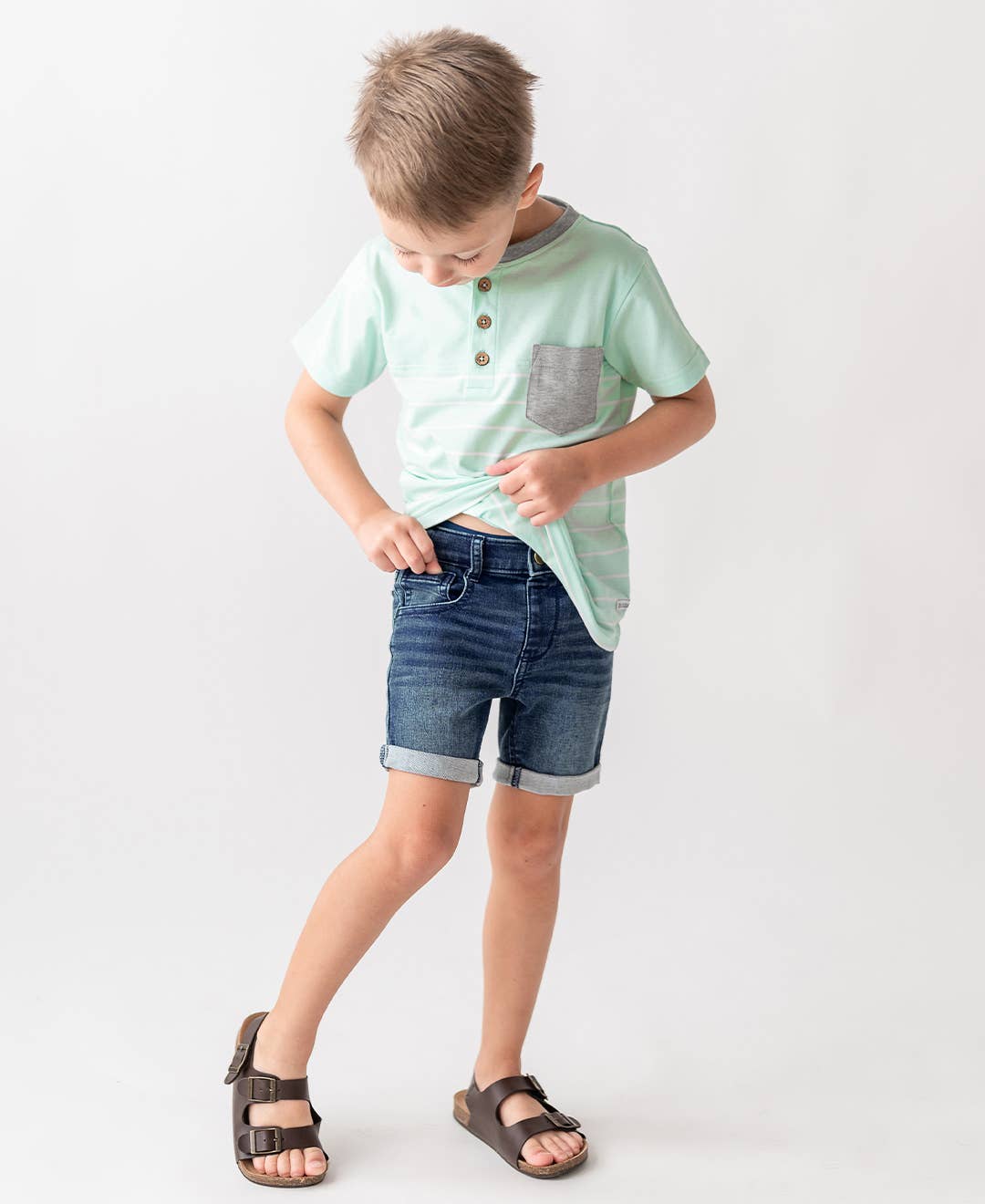 Child wearing a light green shirt with a pocket and blue denim shorts on a white background