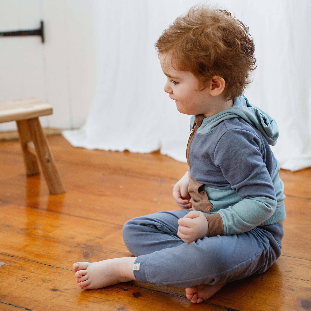 Child sitting on a wooden floor wearing a blue outfit with suspenders.
