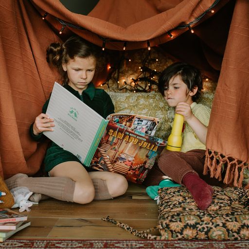 Two children sitting inside a cozy tent reading a book.