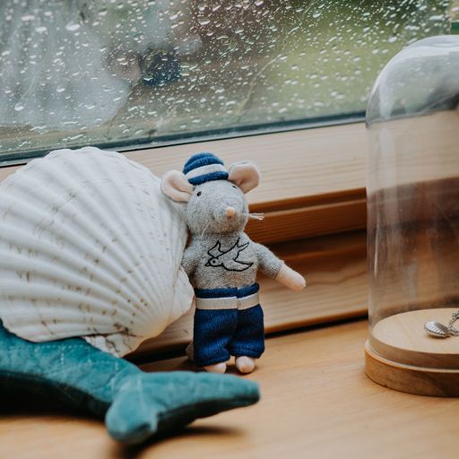 Small toy mouse in a sailor outfit next to a large shell and a glass dome on a wooden surface.