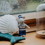 Small toy mouse in a sailor outfit next to a large shell and a glass dome on a wooden surface.