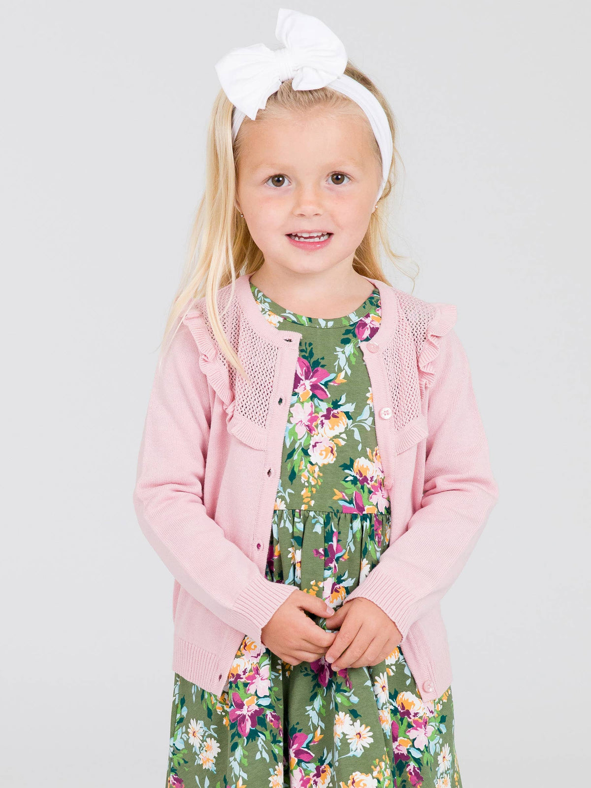 Young girl wearing a pink cardigan over a floral dress on a light gray background