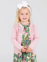Young girl wearing a pink cardigan over a floral dress on a light gray background