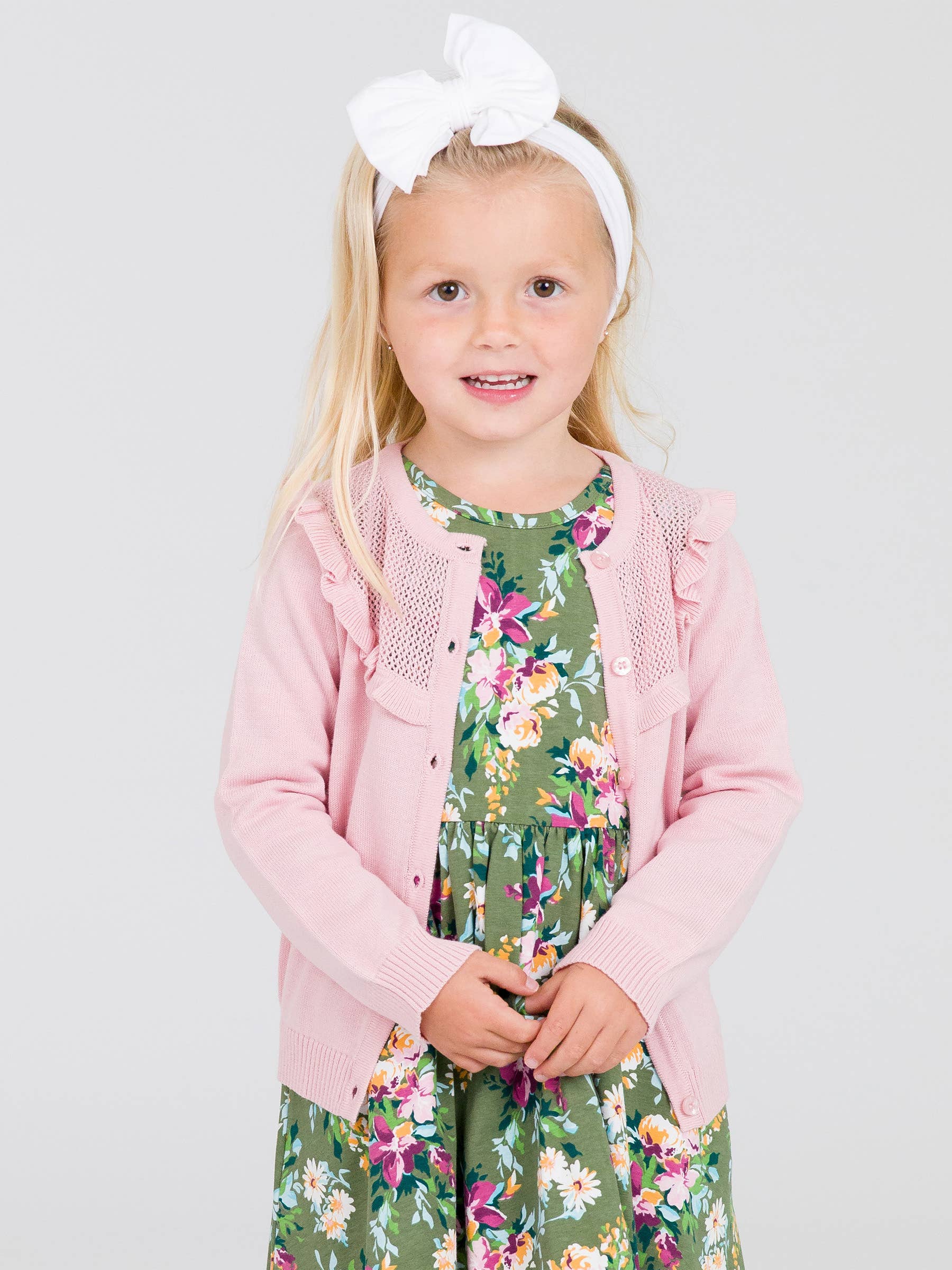 Young girl wearing a pink cardigan over a floral dress on a light gray background