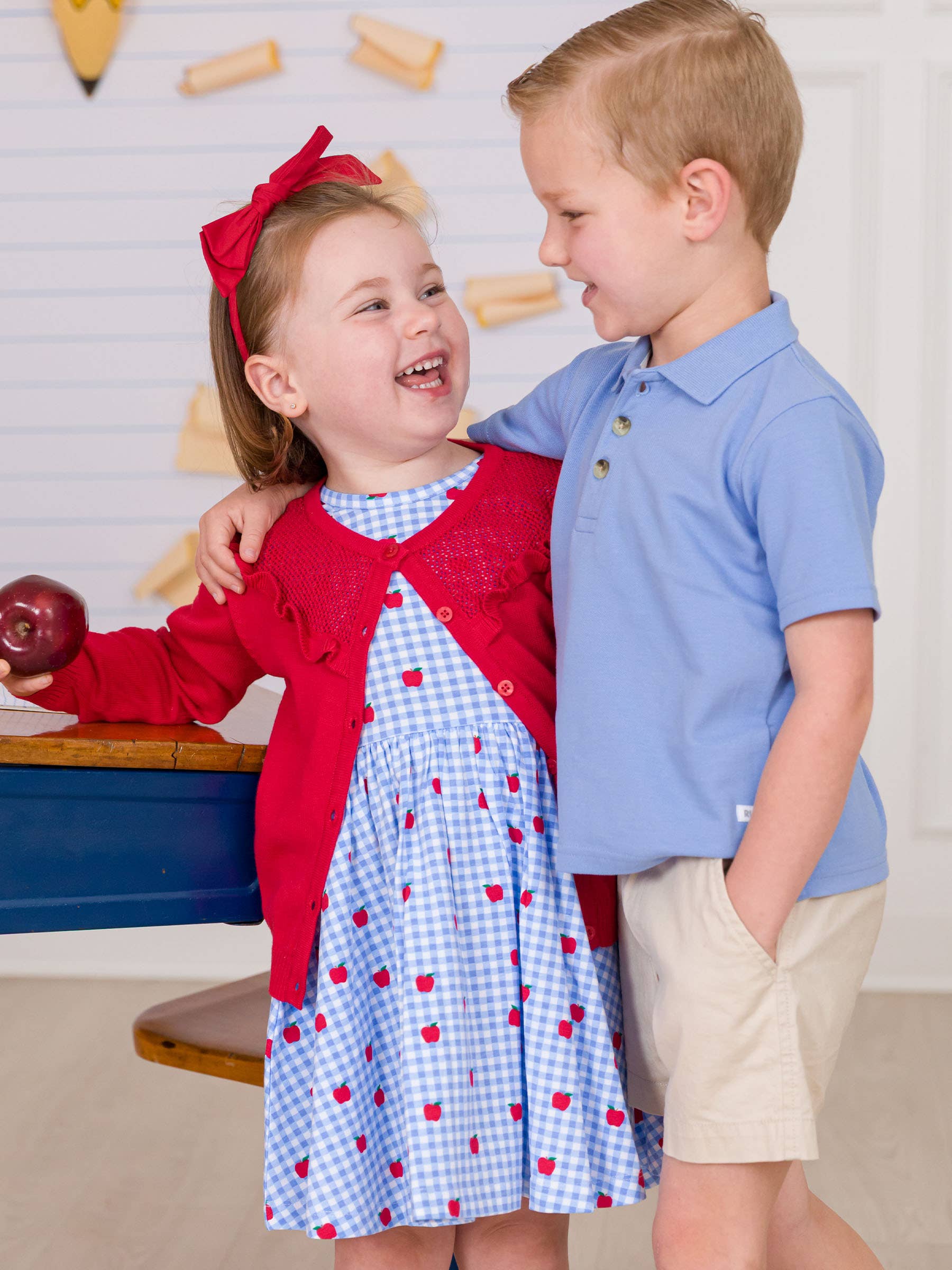Two children, a girl in a red sweater and blue dress with apple pattern and a boy in a blue shirt and beige shorts, standing close together.