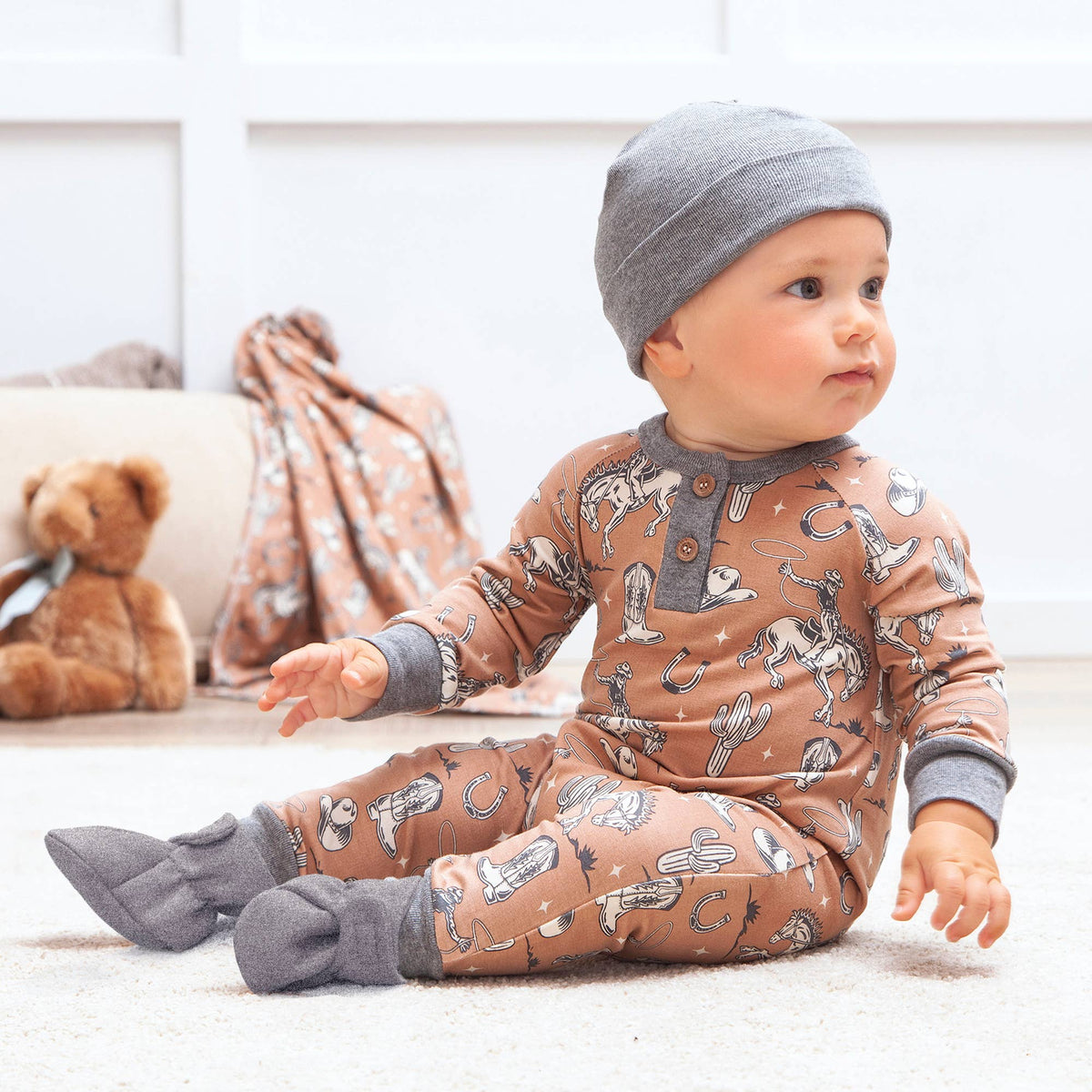 Baby wearing a brown onesie with animal patterns and gray hat and socks, sitting on a white floor.