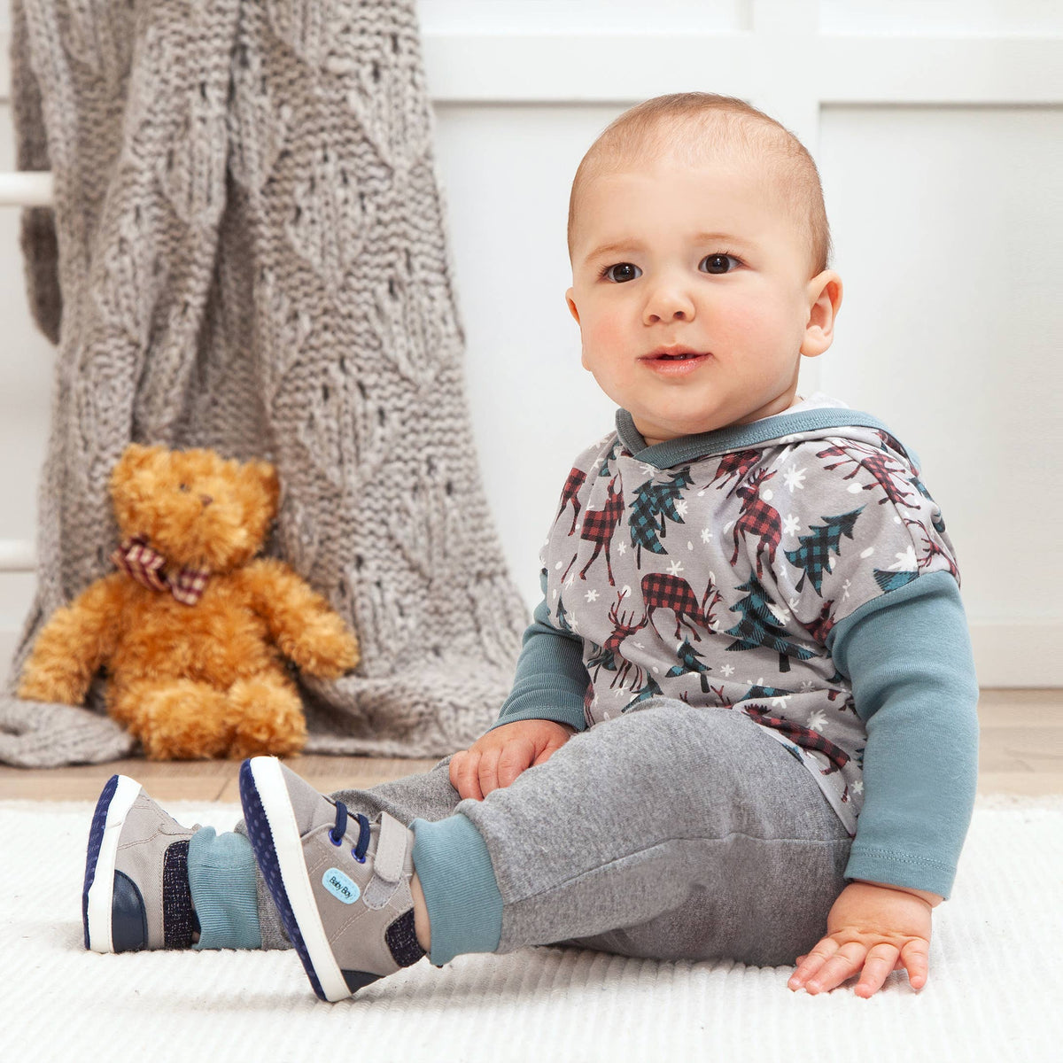 Baby sitting on a white surface with a teddy bear and knitted blanket in the background.