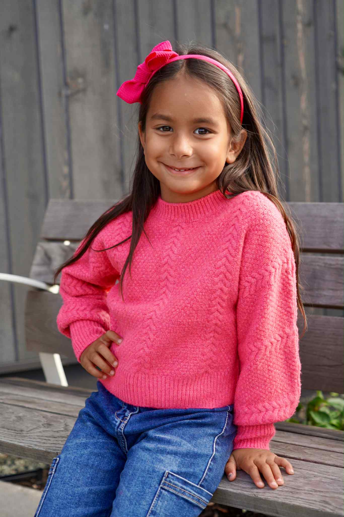 Young girl wearing a pink sweater and blue jeans sitting on a wooden bench.