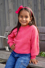 Young girl wearing a pink sweater and blue jeans sitting on a wooden bench.