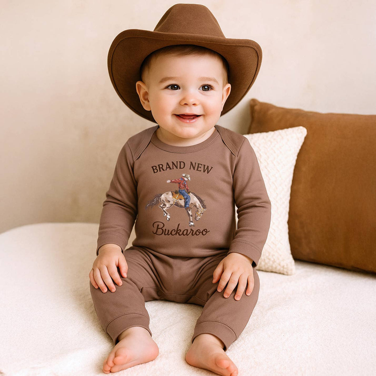 Baby wearing a brown cowboy hat and 'Brand New Buckaroo' outfit sitting on a couch.