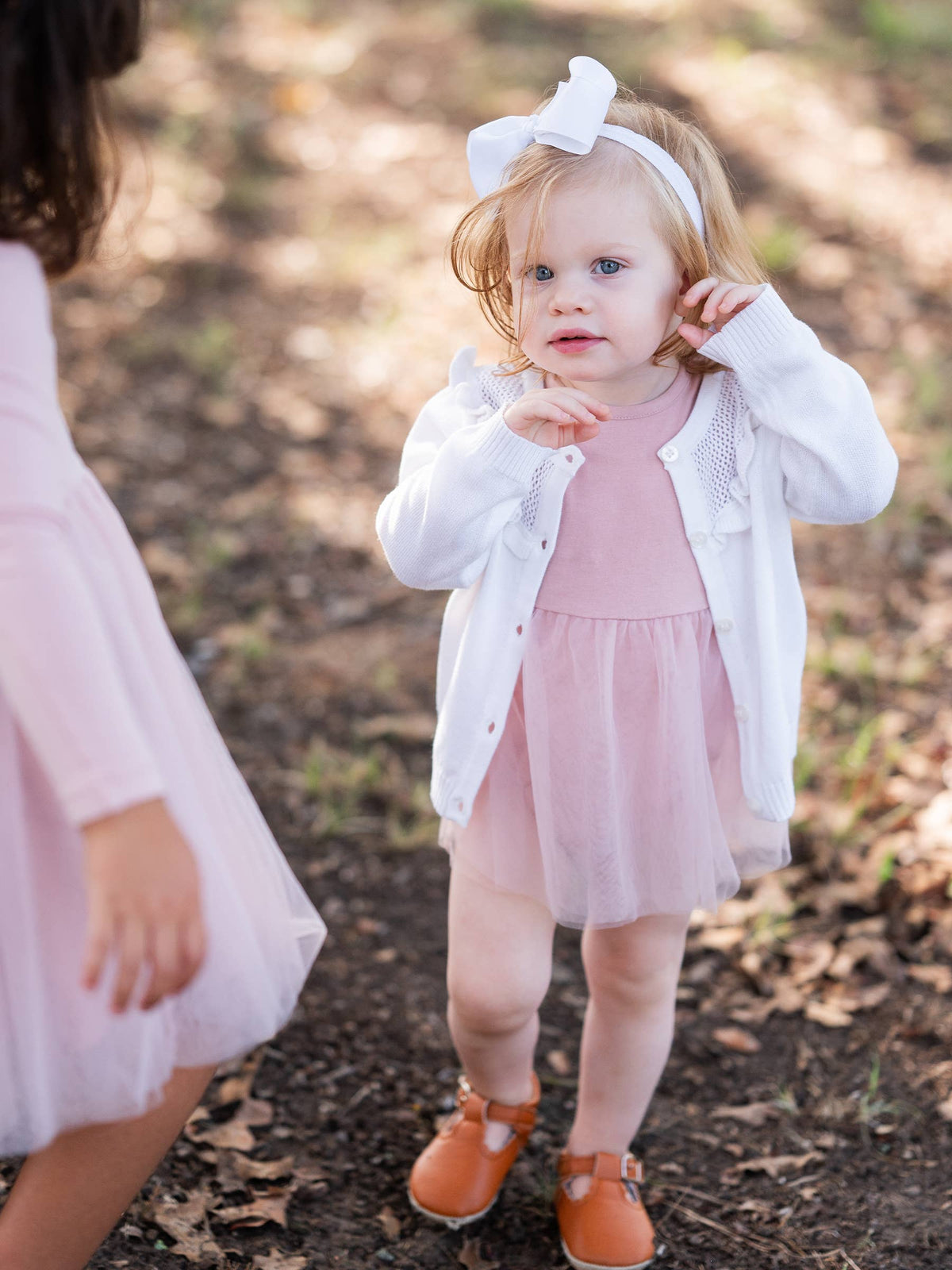 Young girl in a pink dress and white cardigan standing outdoors on a leafy ground.