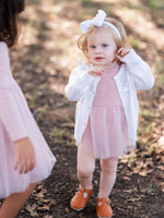 Young girl in a pink dress and white cardigan standing outdoors on a leafy ground.