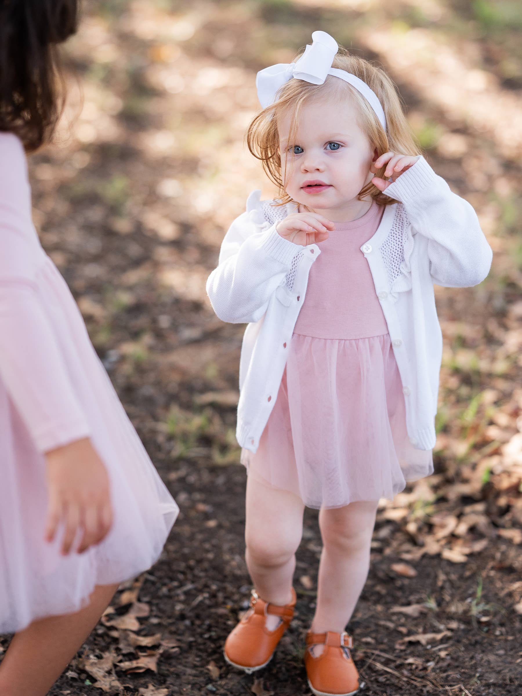 Young girl in a pink dress and white cardigan standing outdoors on a leafy ground.