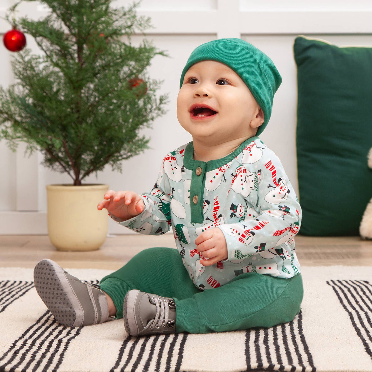 Baby in green hat and matching outfit sitting on a striped rug with a Christmas tree in the background.