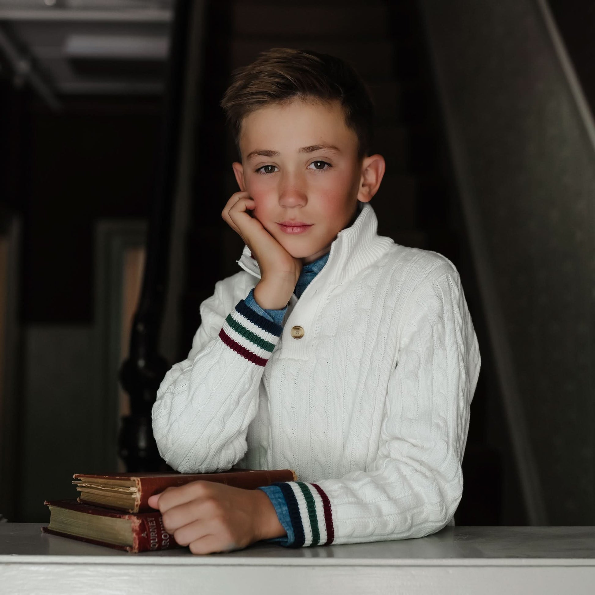 Young boy in a white cable knit sweater with striped cuffs, sitting at a table with books.