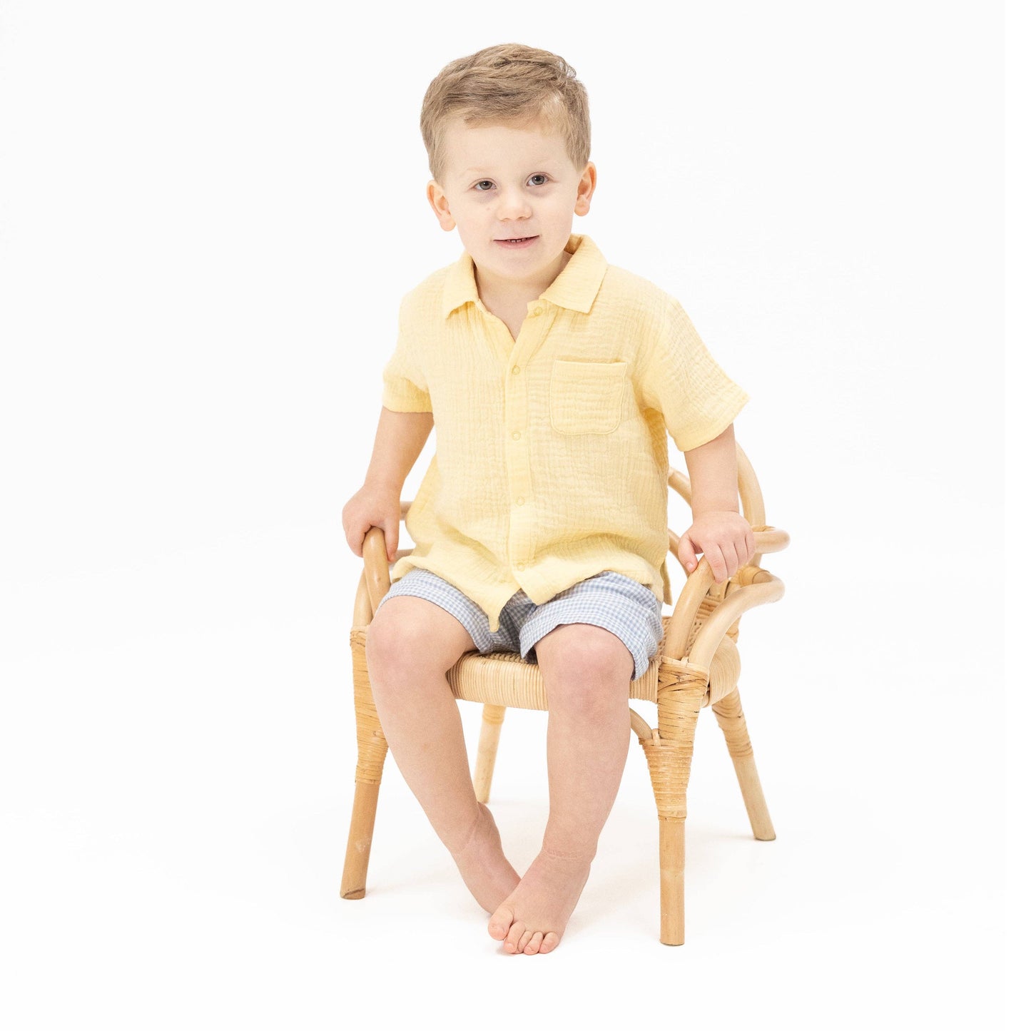 Child sitting on a small chair wearing a yellow shirt and striped shorts against a white background