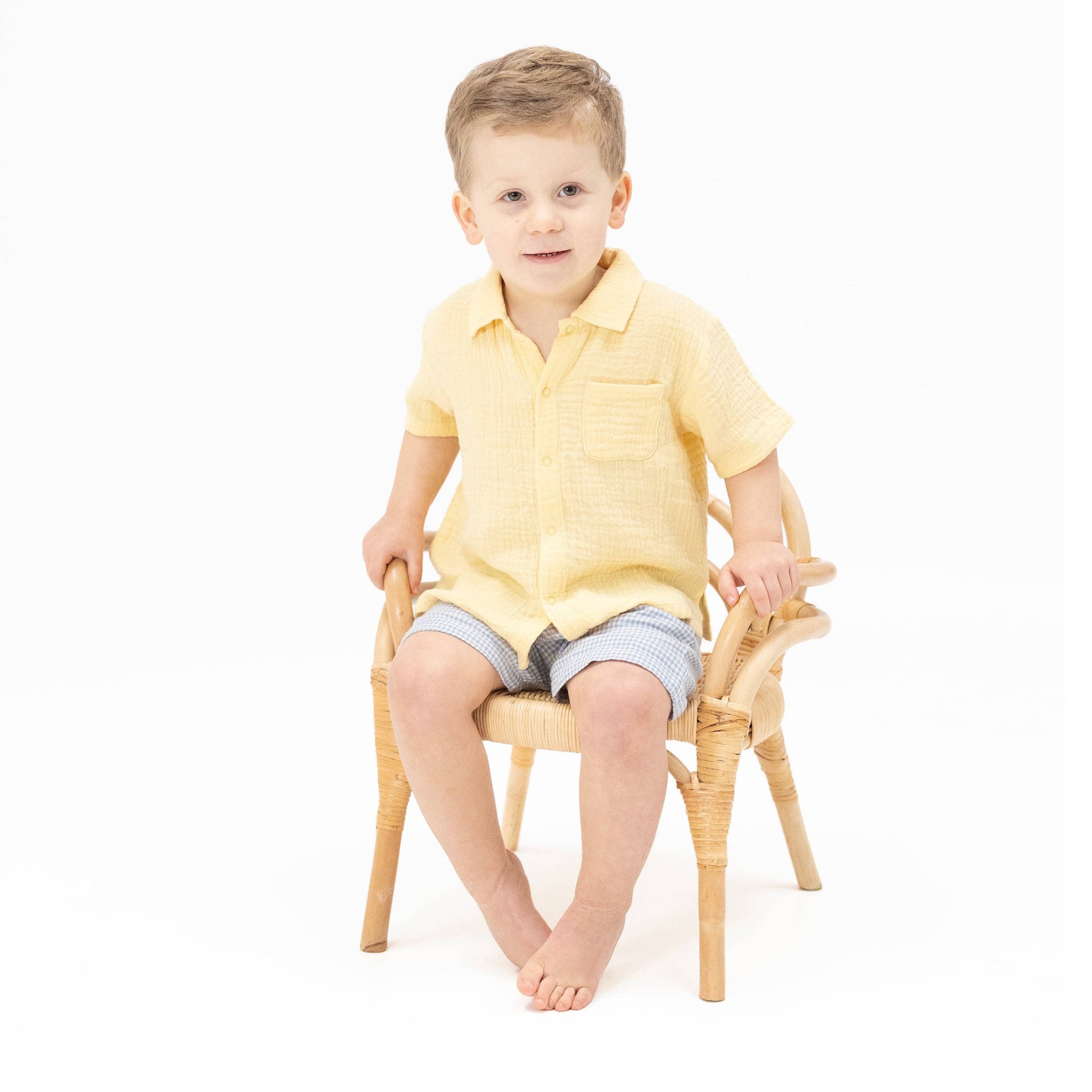 Child sitting on a small chair wearing a yellow shirt and striped shorts against a white background