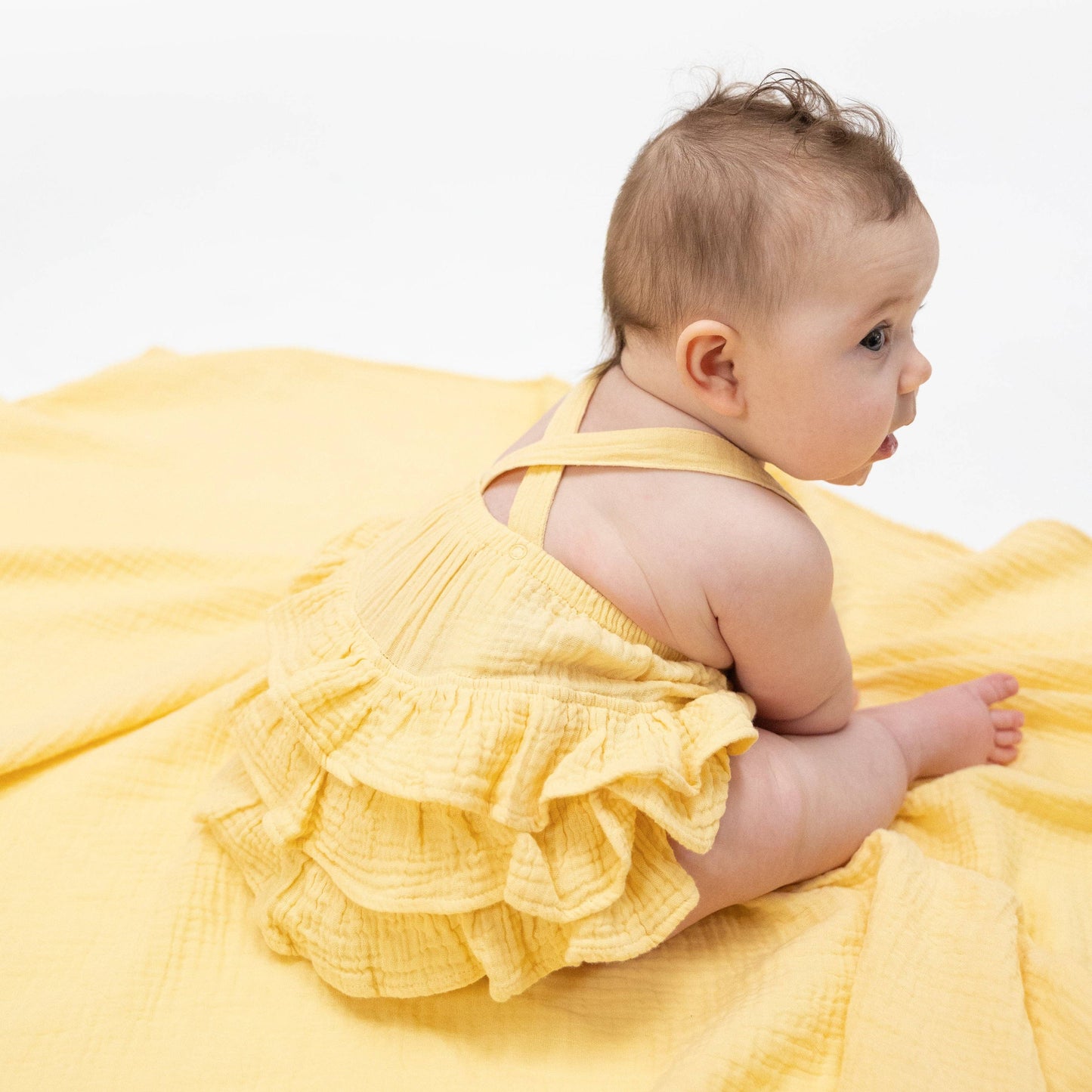 Baby in a yellow sunsuit sitting on a yellow blanket against a white background