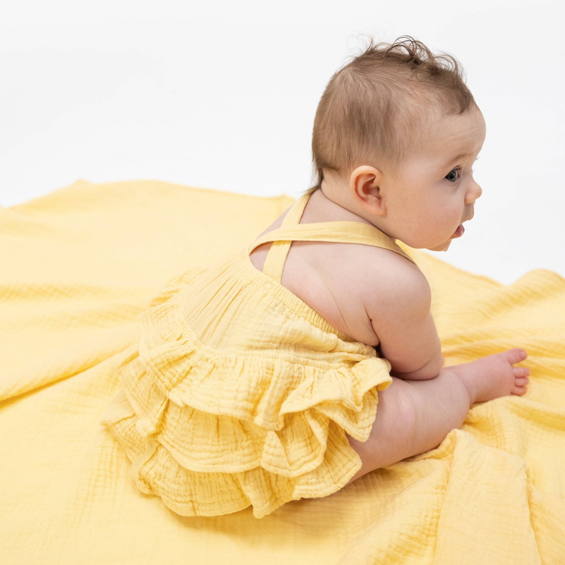 Baby in a yellow sunsuit sitting on a yellow blanket against a white background