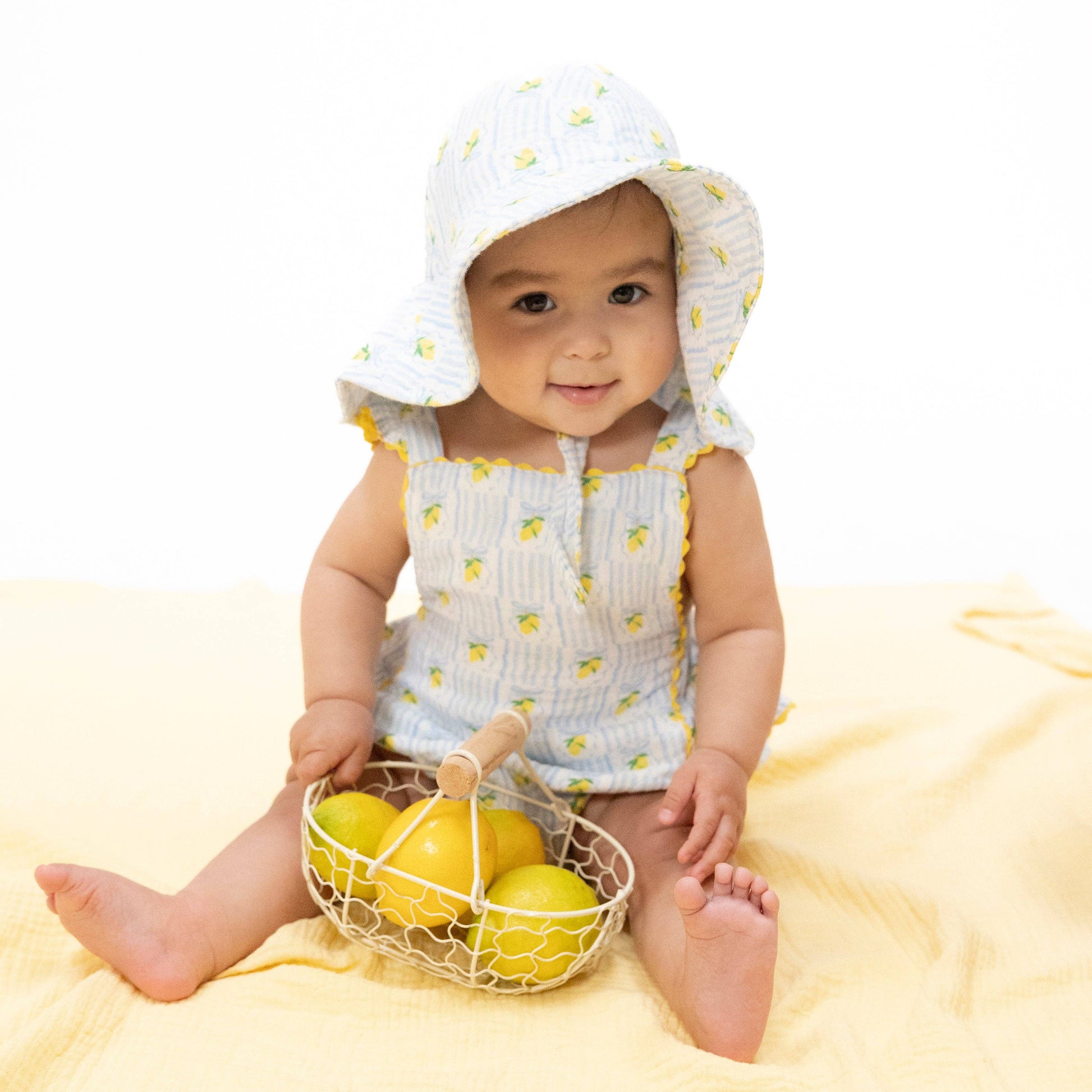 Baby in a lemon-themed outfit holding a basket of lemons on a yellow blanket.