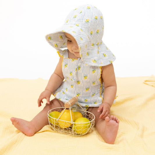 Baby wearing a lemon-patterned outfit with a matching hat, sitting on a yellow blanket with a basket of lemons.