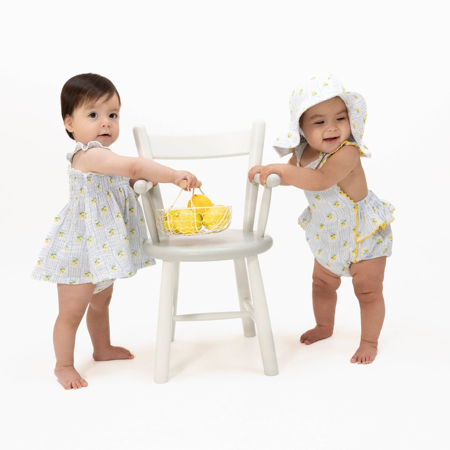 Two young children in matching outfits with a white chair and yellow basket on a white background