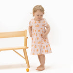 Child wearing a dress with peach patterns standing next to a wooden chair on a white background