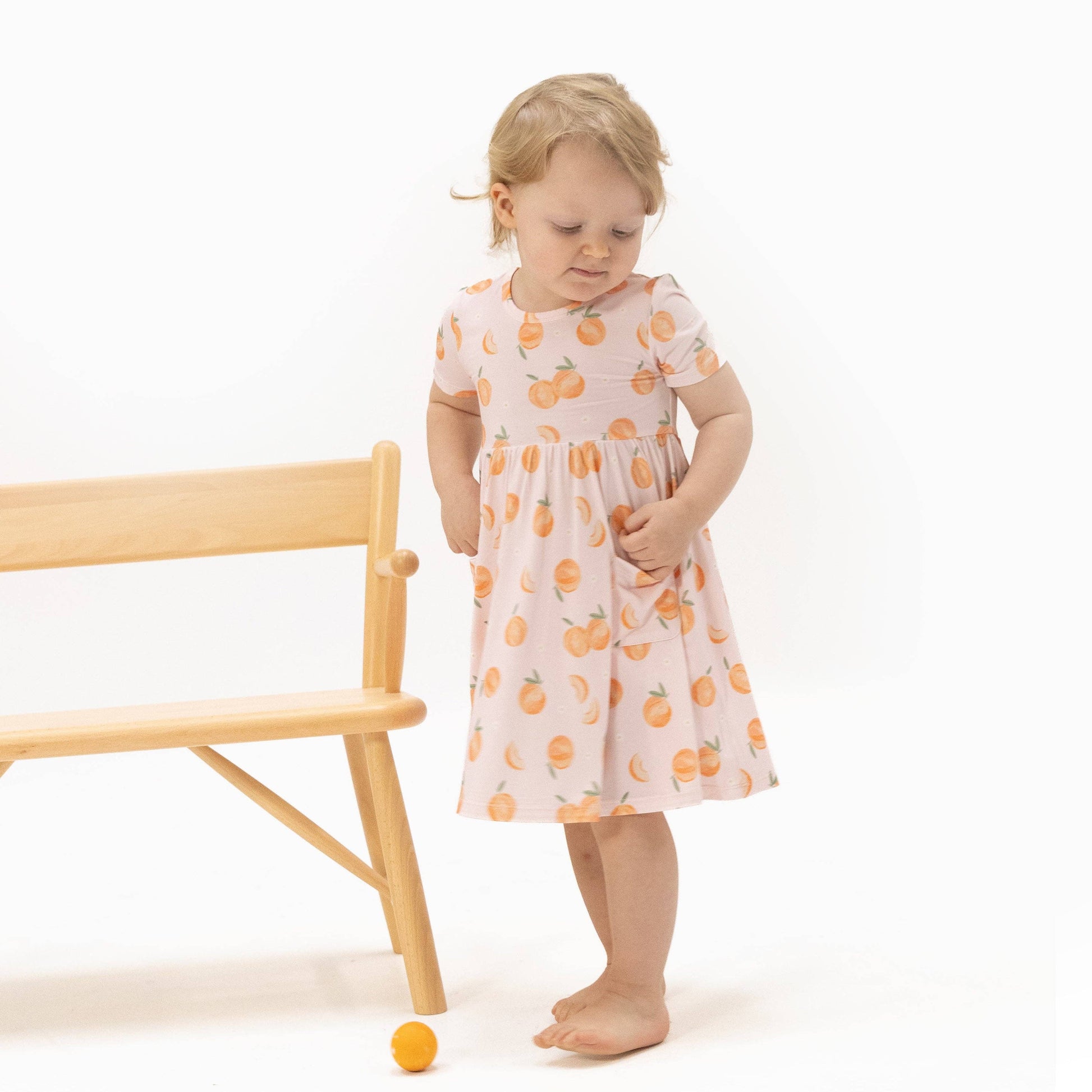 Child wearing a dress with peach patterns standing next to a wooden chair on a white background