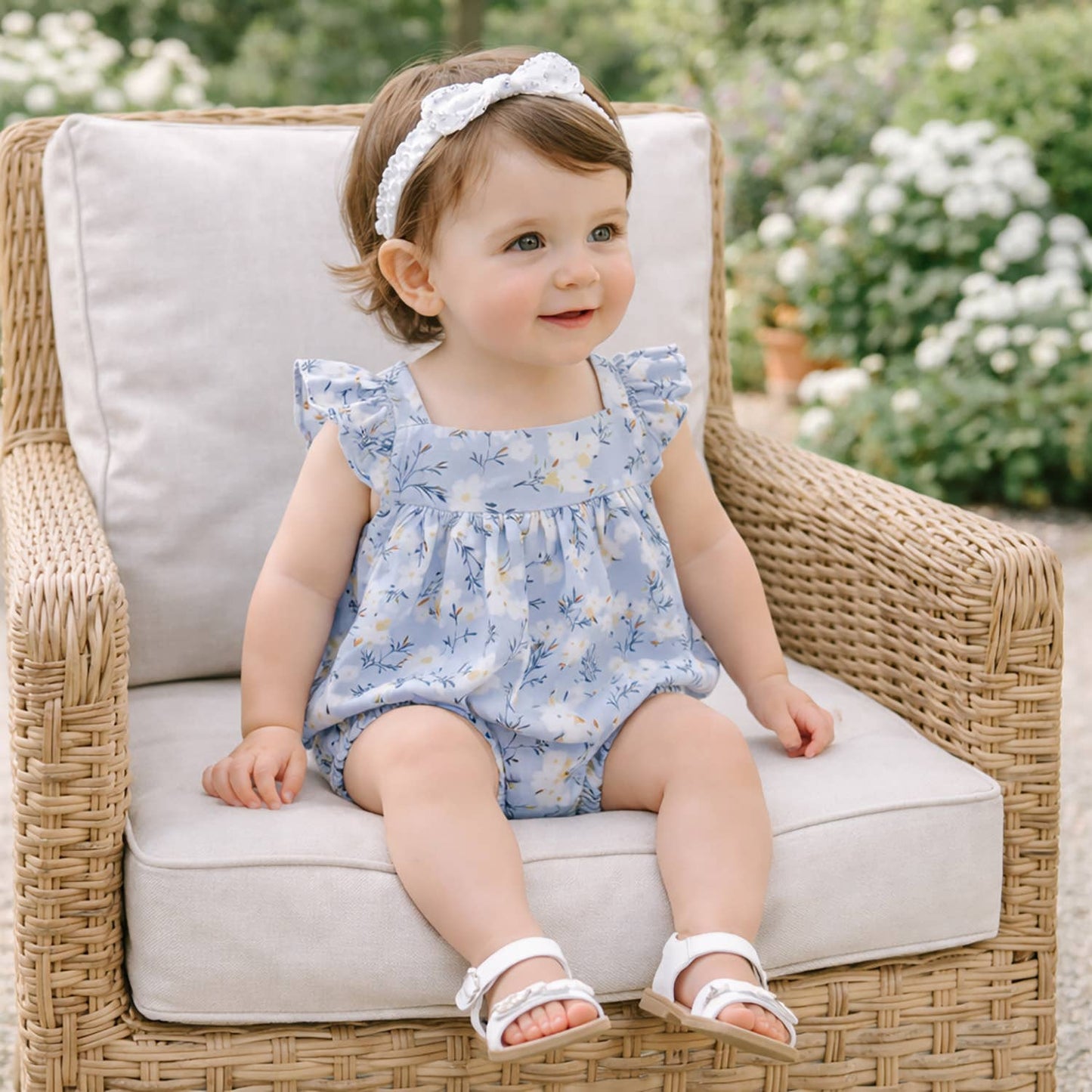 Baby in a floral romper sitting on a wicker chair outdoors.