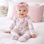 Baby wearing a floral onesie and pink headband sitting on a bed.