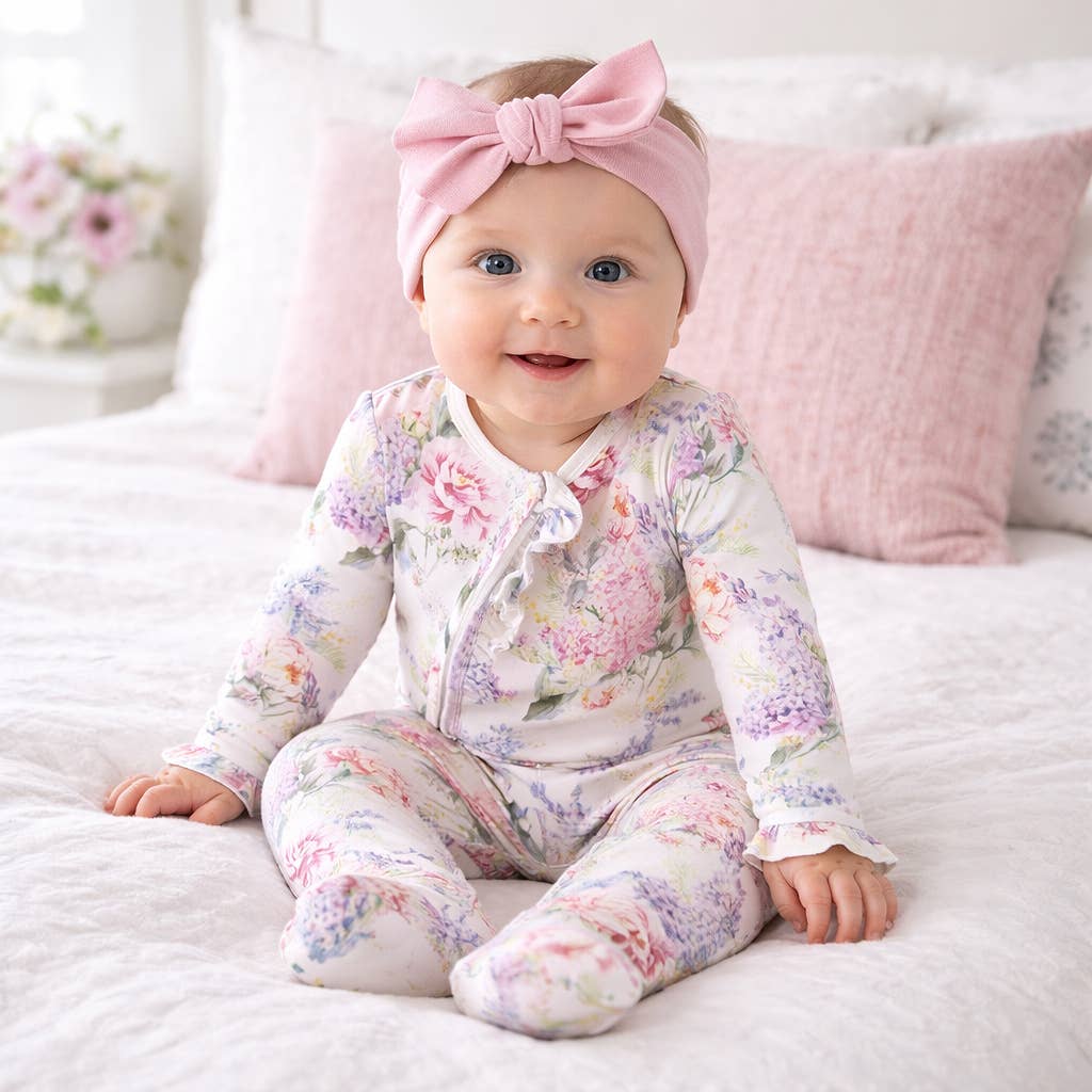 Baby wearing a floral onesie and pink headband sitting on a bed.