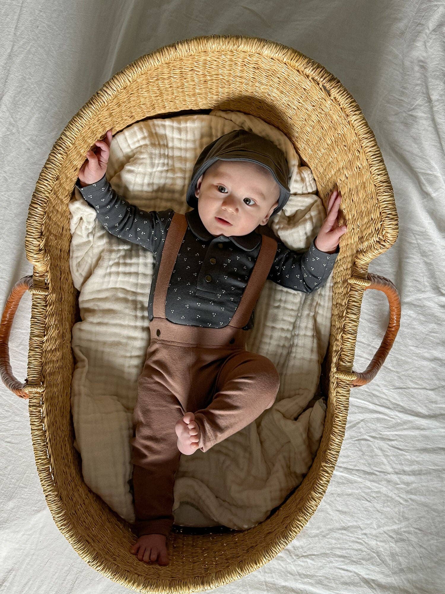 Baby in a moses basket wearing brown pants with suspenders on a neutral background