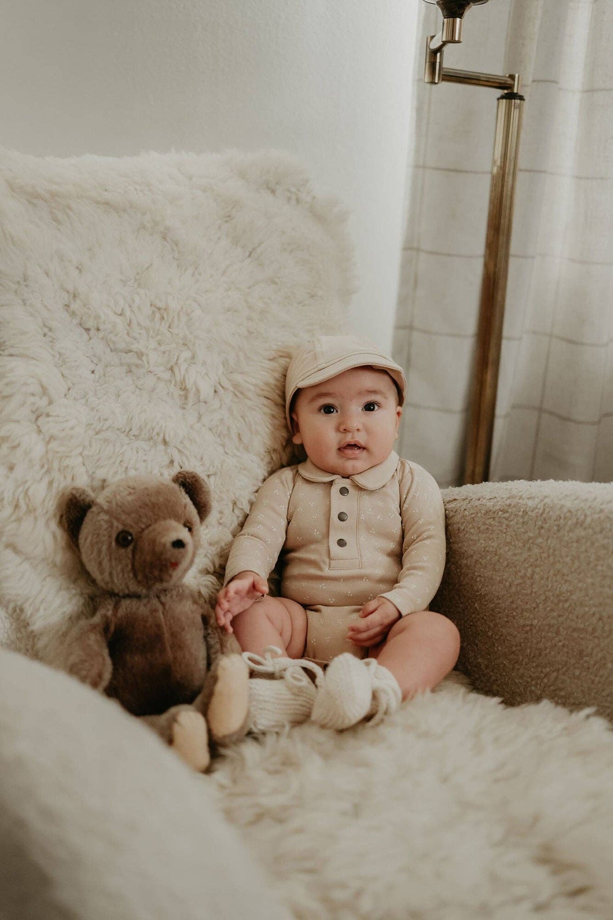 Baby in a beige outfit sitting on a fluffy chair with a teddy bear