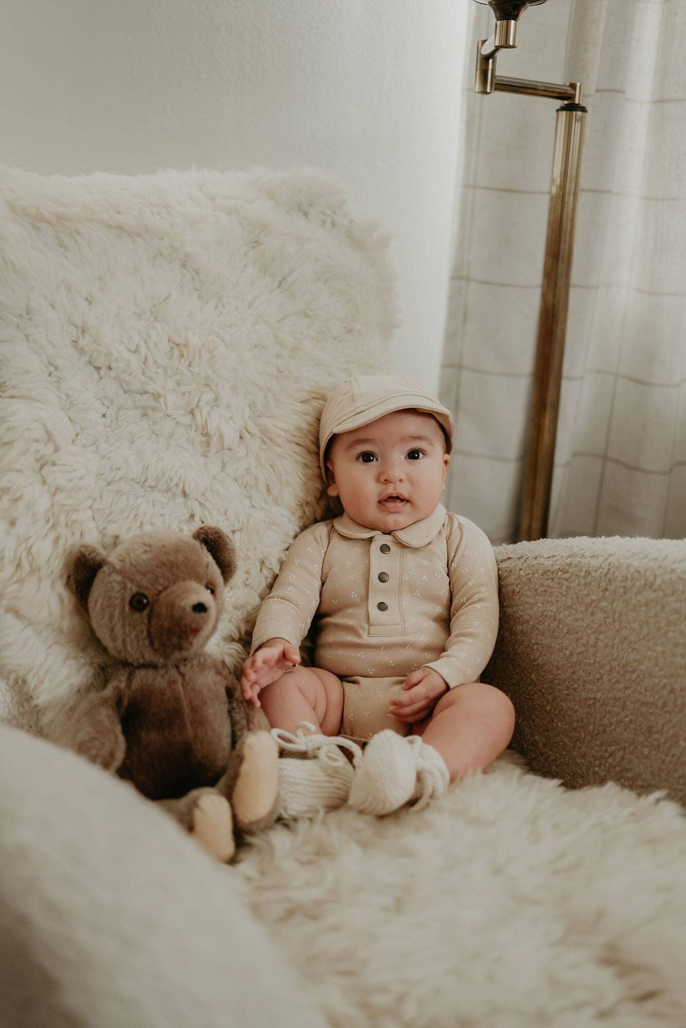 Baby in a beige outfit sitting on a fluffy chair with a teddy bear