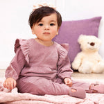 Baby in a mauve outfit sitting on a bed with a teddy bear and purple pillow in the background