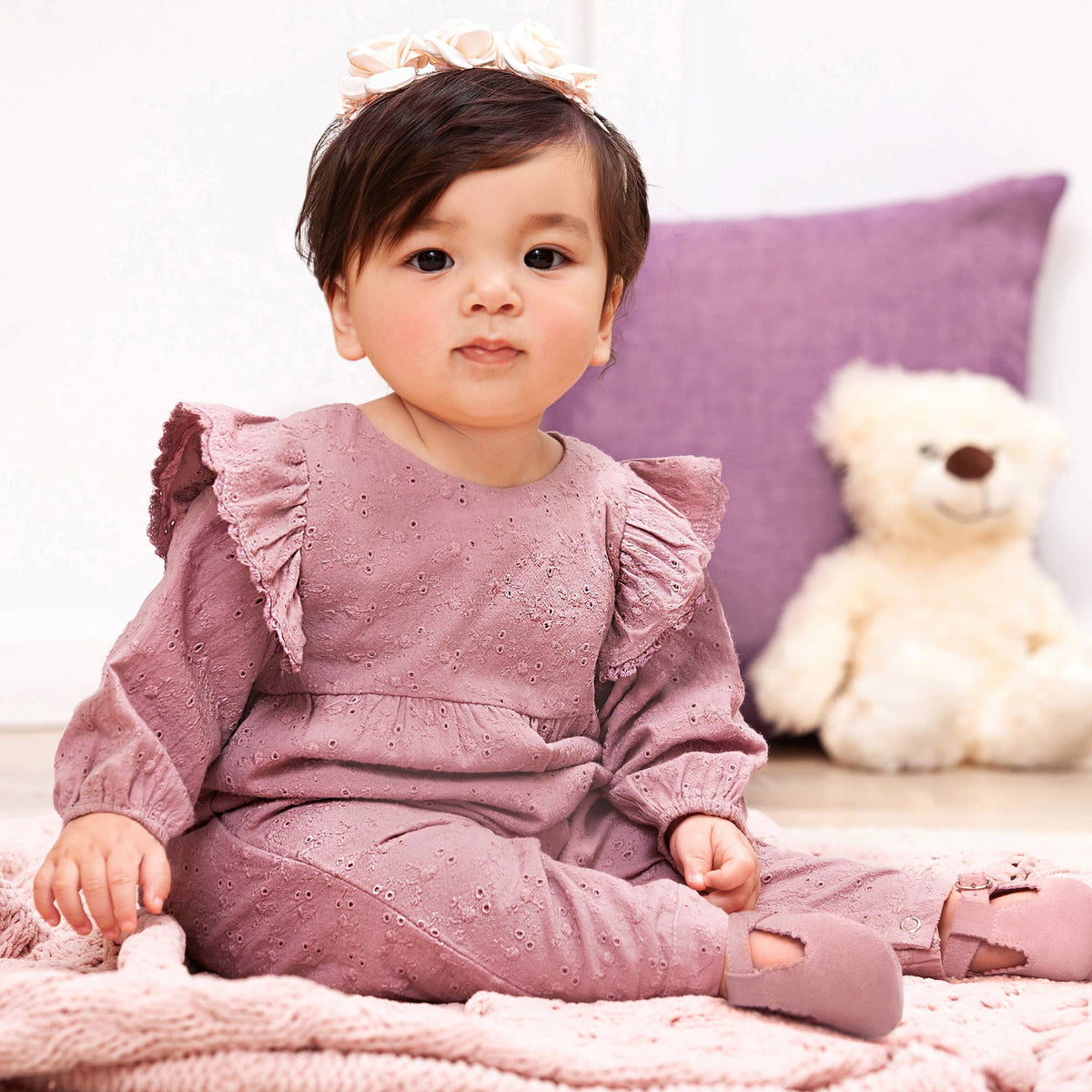 Baby in a mauve outfit sitting on a bed with a teddy bear and purple pillow in the background