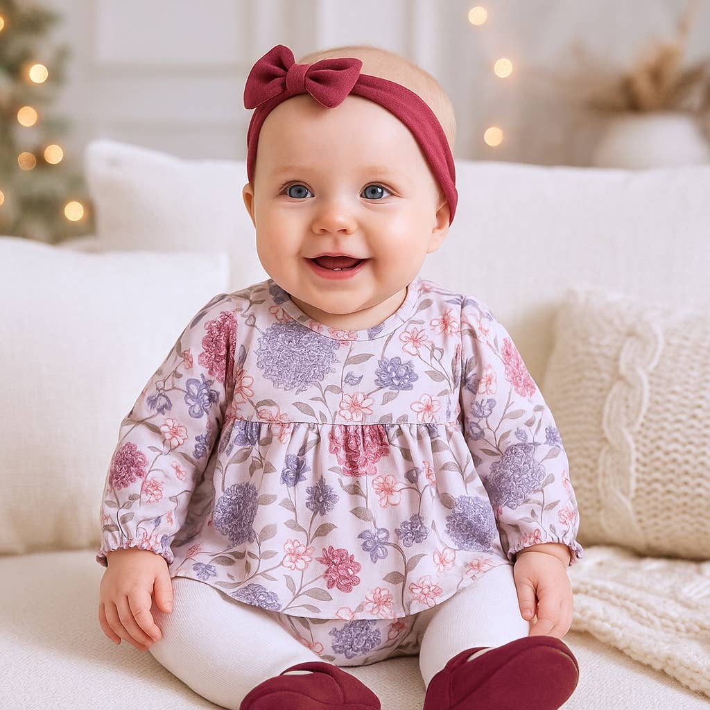 Baby wearing a floral top and red headband sitting on a couch with a Christmas tree in the background.