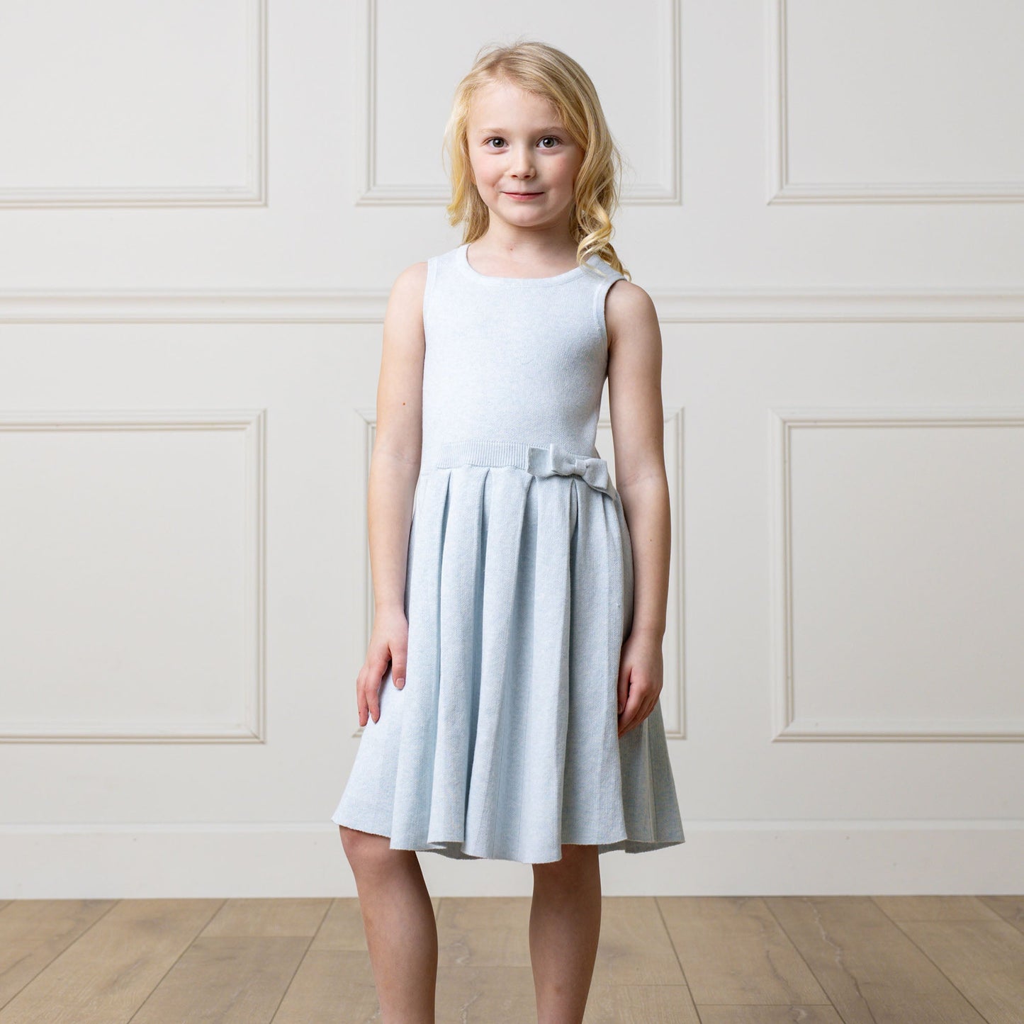 Young girl wearing a light blue dress against a white paneled wall.